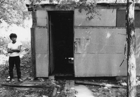 a boy standing beside the shower facilities at a migrant workers village. the facility has no visible door, metal sides and no lighting.