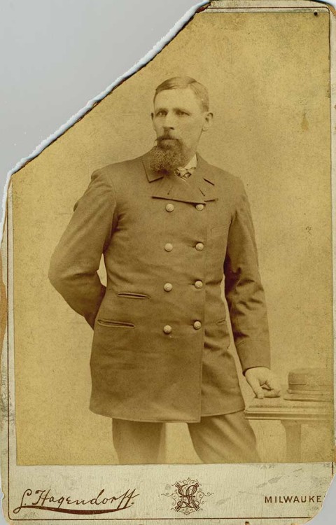 A sepia-toned portrait of a man standing with his arm behind his back.