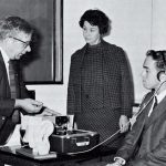 A black and white image of three people testing an audiometer, one man has headphones on his ears while a woman looks on and another man is testing a read-out from the machine.