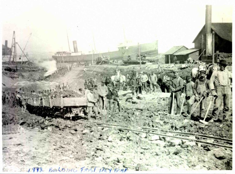 A black and white images showing workers at the American Steel Barge Company. They are in a muddy field beside a track leading to the docks