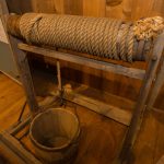 A color photograph of a windlass with a rope wound around its beam and a wooden bucket attached to the line below.